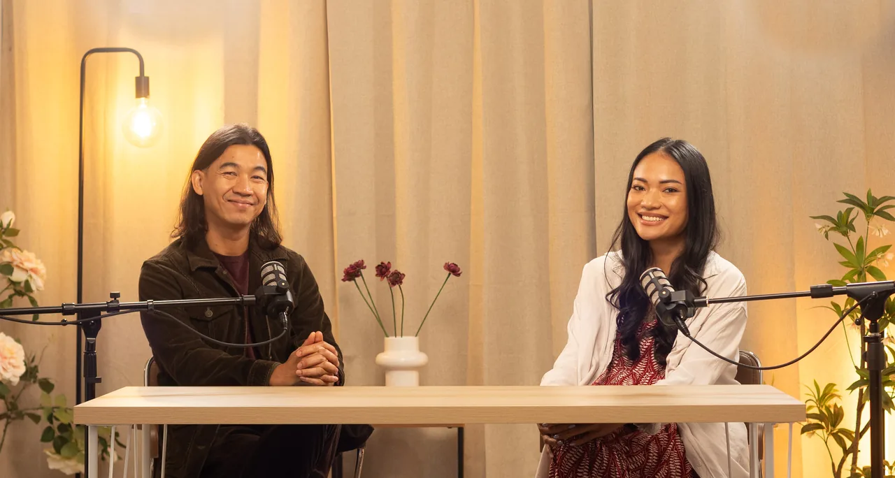 Two podcast hosts smiling at a desk with microphones under warm studio lights